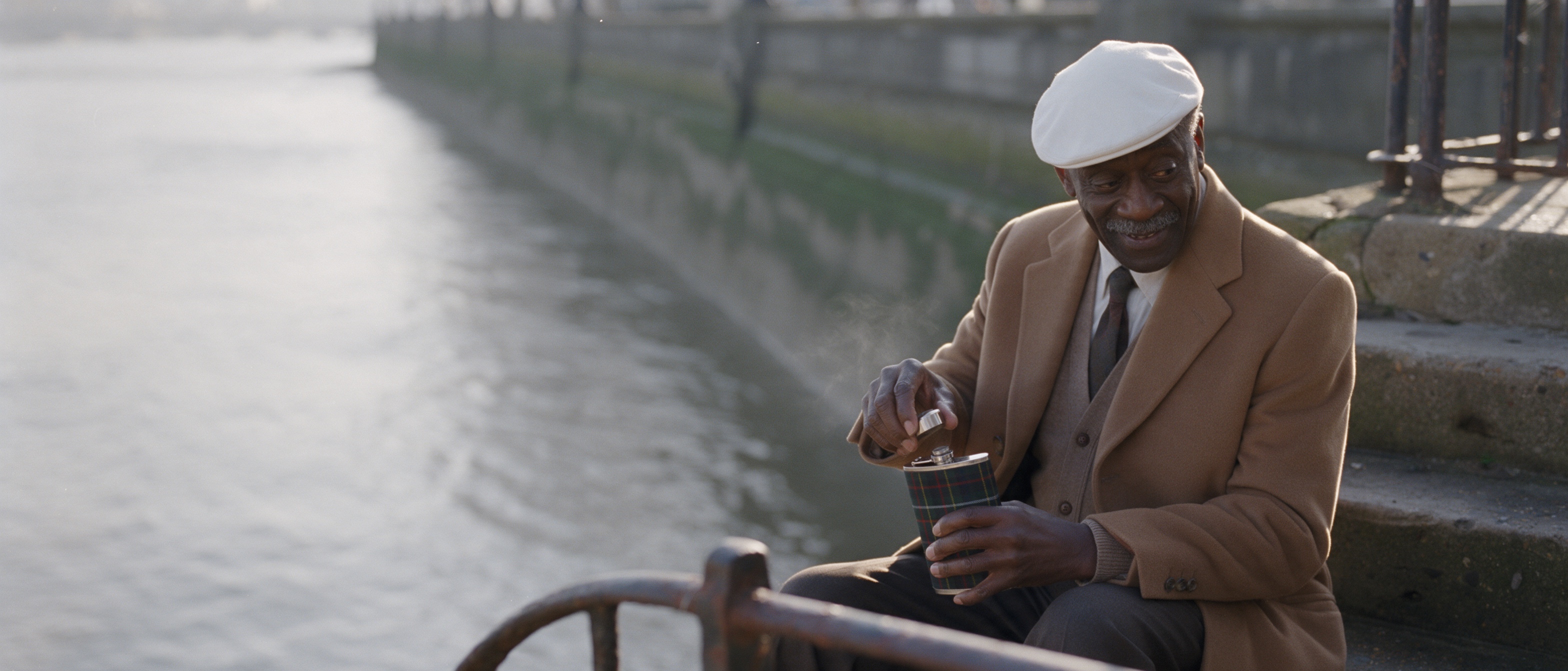 An old man in a beret and camel coat sitting by a river