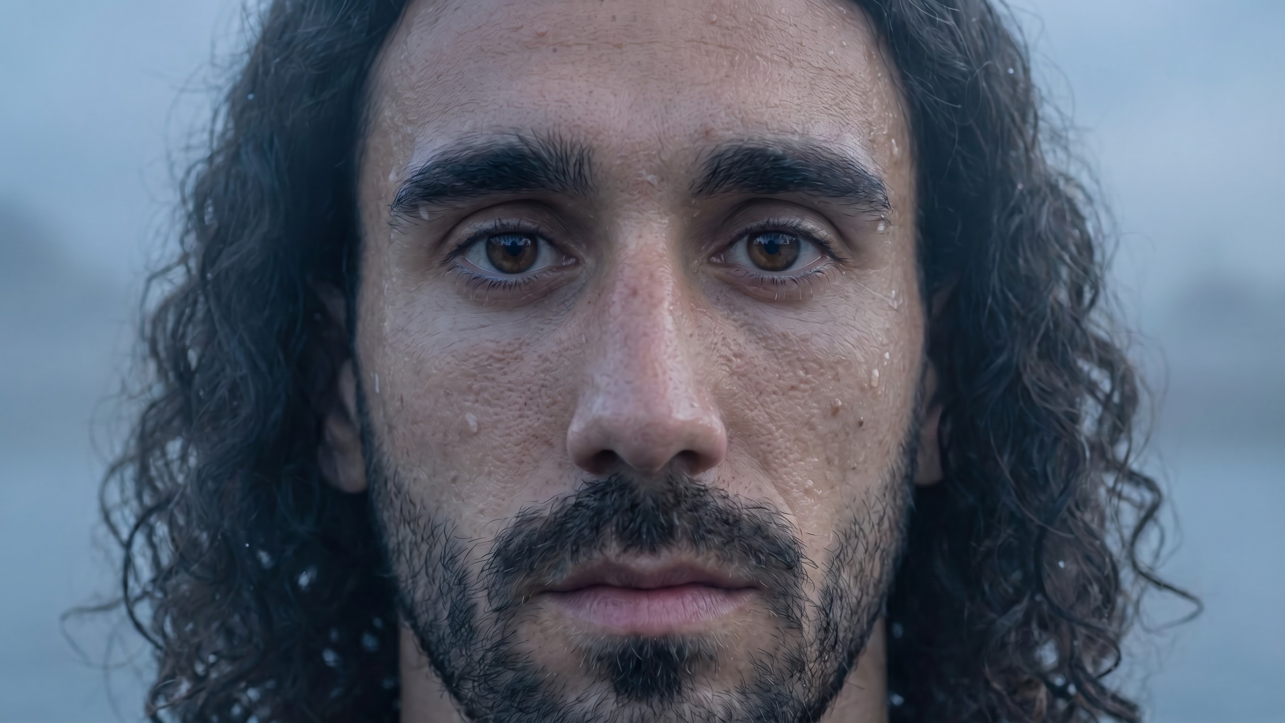 A frontal close-up of Marc Cucurella soaked with rain, hair loose, against a pale grey river sky