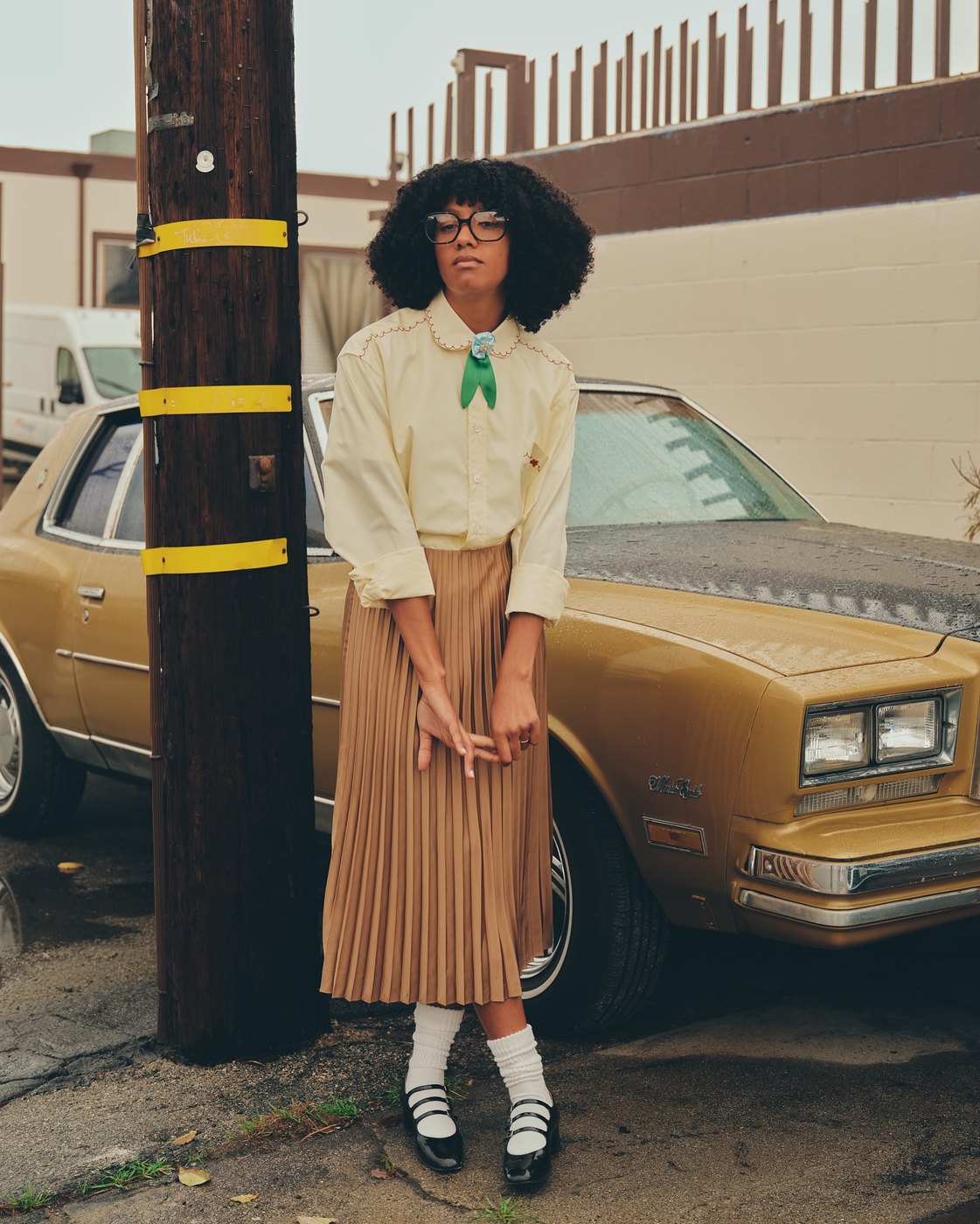 A model in a scalloped collar shirt, pleated skirt, white socks and loafers beside a car