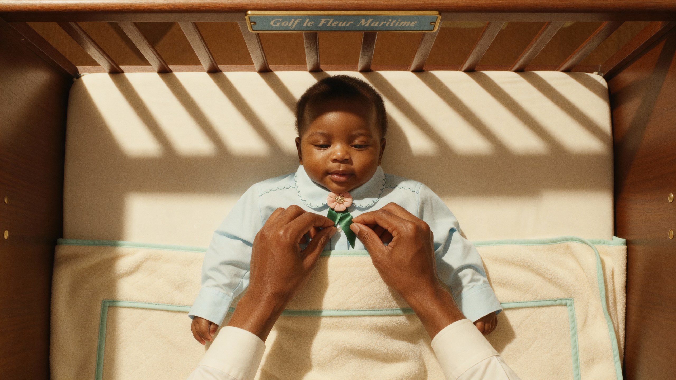 Hands tying a green bow at the collar of a baby in the same pale blue shirt, inside a cot marked Golf le Fleur Maritime