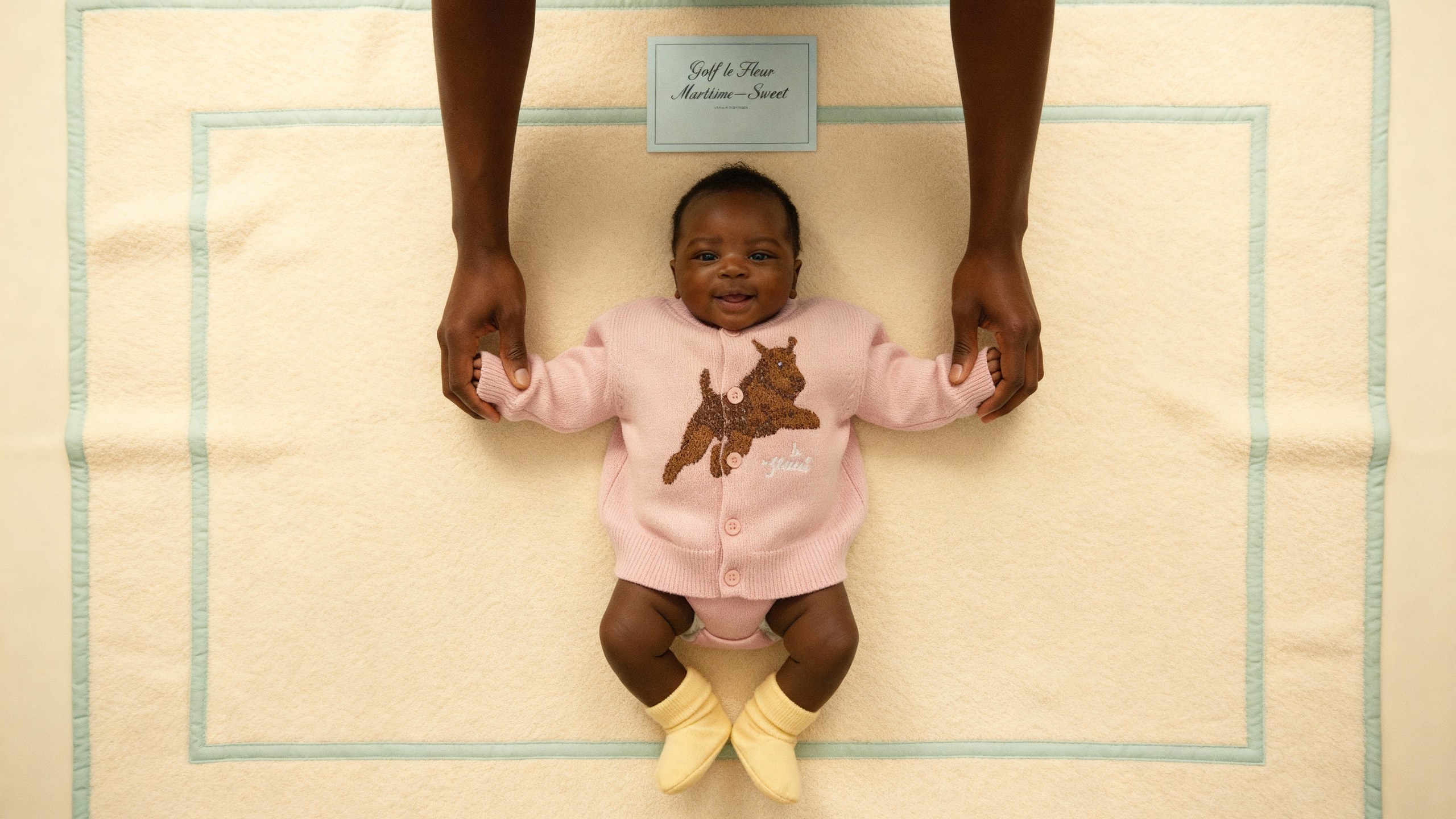 A baby in a pink babygrow carrying the same terrier, photographed from directly above on a blanket