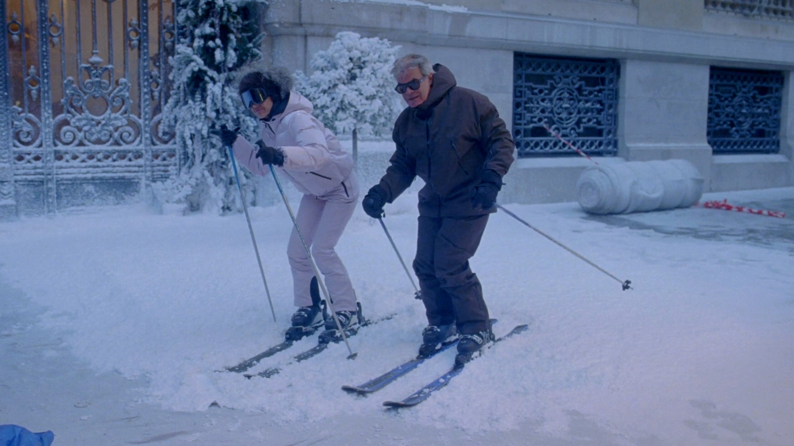 The reference frame filmed by the production: a woman in a pale pink ski suit with a fur-trimmed hood and mirrored goggles and a man in a brown ski suit and sunglasses, on skis beside an iron gate