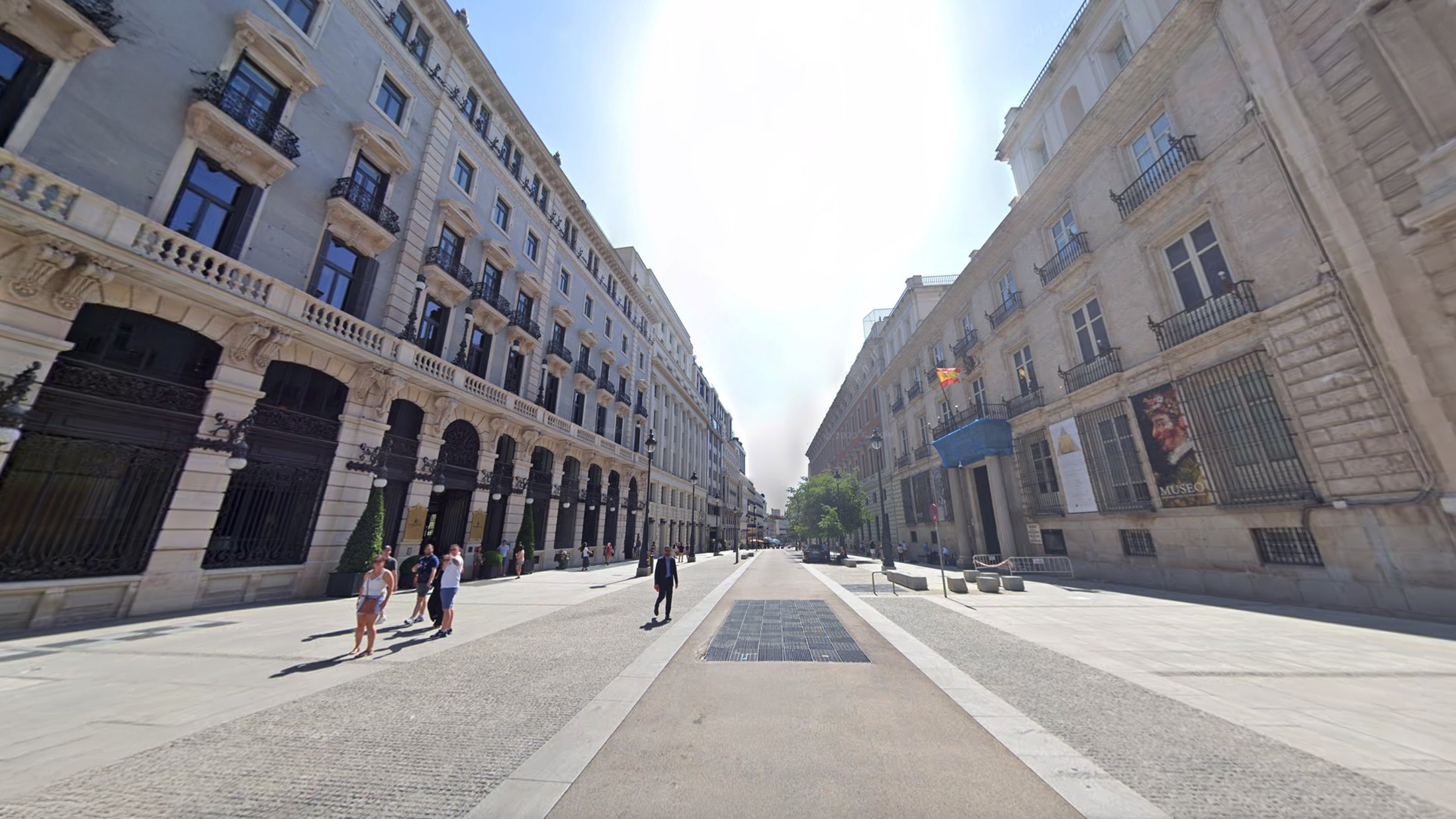 A Madrid street on an ordinary day, an arcade of arches down the left side, a flag and an exhibition poster on the building opposite