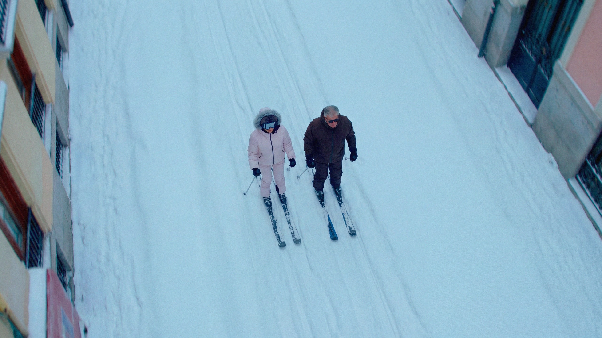 A drone shot looking straight down a snow-covered Madrid avenue, two skiers leaving parallel tracks between the buildings