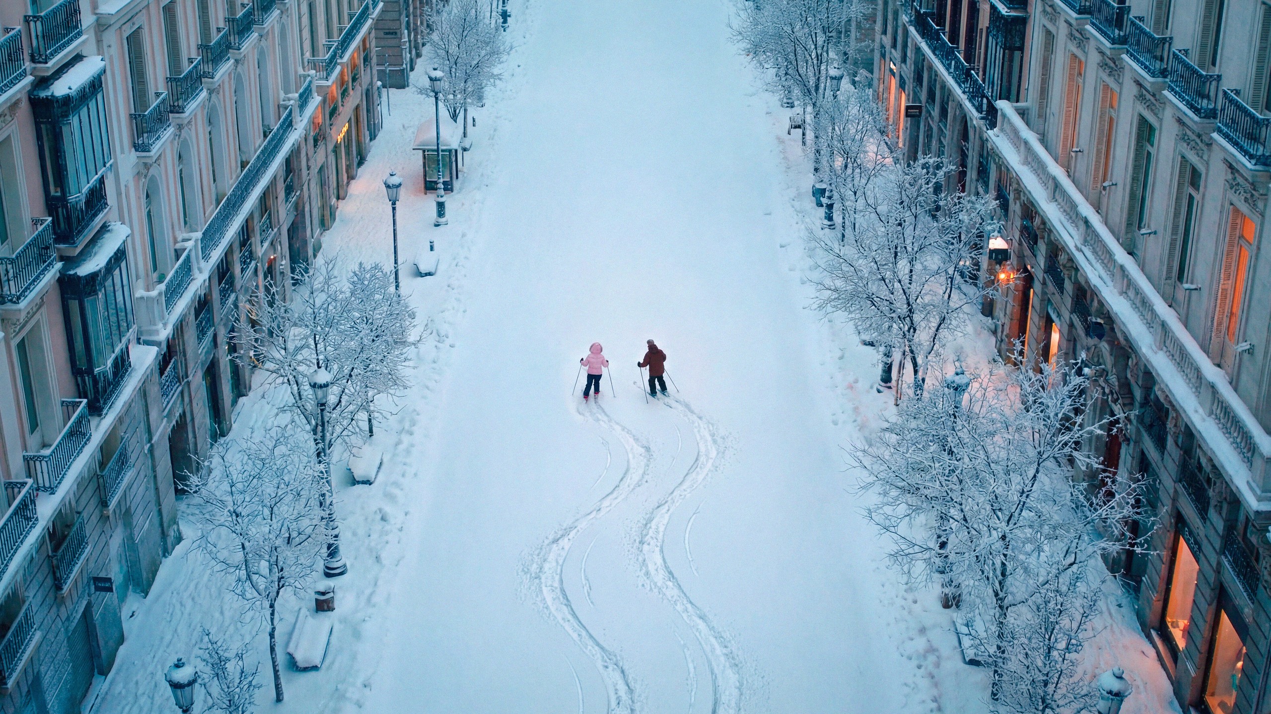 A wide snow-covered Madrid avenue at dusk with lit shopfronts, snowed trees and two distant skiers
