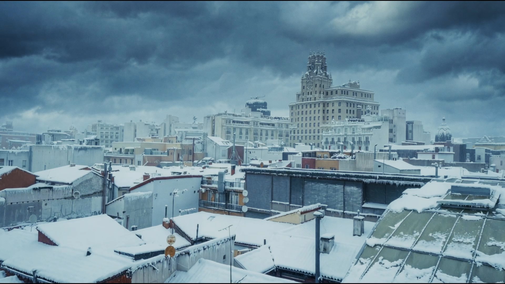 Madrid rooftops under heavy snow beneath a dark storm sky, the Telefónica tower on Gran Vía in the distance